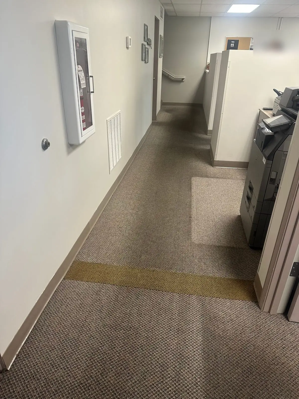 An office hallway at Useless Bay Golf & Country Club before cleaning, with a dark traffic lane worn down the middle of the grey loop carpet and one lighter cleaned patch beside the copier