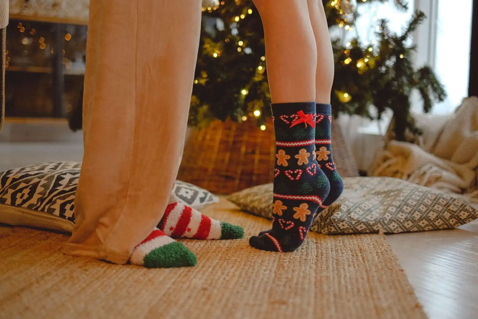 Two people in festive socks standing on a woven jute rug in front of a lit Christmas tree